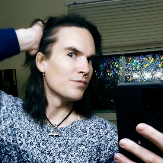 a mirror selfie of Dune, a white trans femme with dark hair. she's wearing a gradient blue knit sweater and holding a hand in her hair, holding it back over her ear.